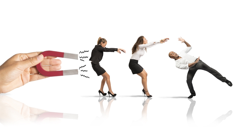 Two women and one man dressed for business are being pulled back by a large hand holding a magnet.
